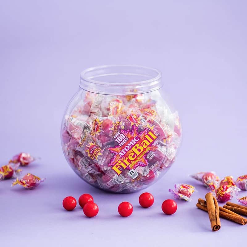 a round container of atomic fireball candies, with some candies and cinnamon sticks scattered around