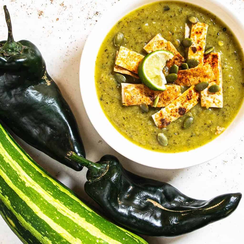 a bowl of green soup surrounded by poblanos and zucchini.