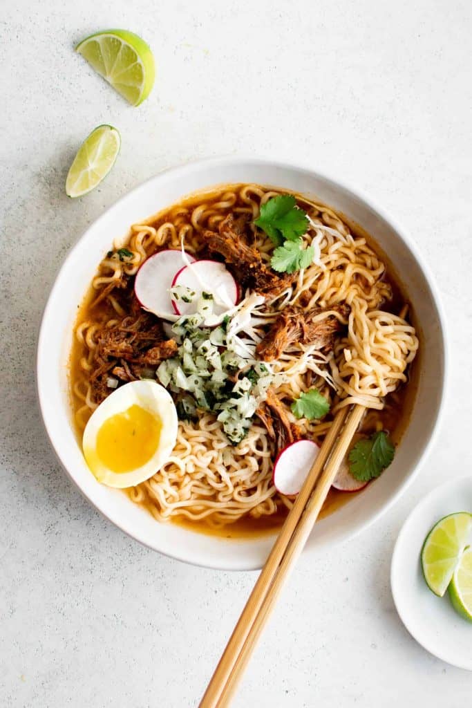 bowl of birria ramen with egg, radish, and lime wedges.