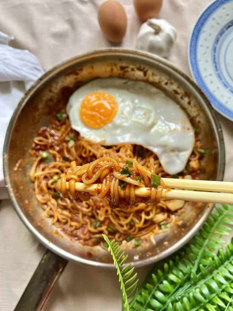 closeup of chopsticks holding ramen noodles with blurred bowl of ramen with fried egg in the background.