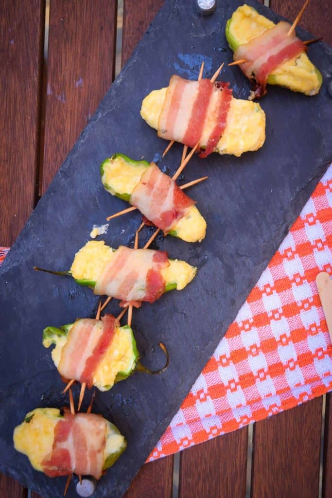 several grilled jalapeño poppers wrapped in bacon on a stone slab dish.