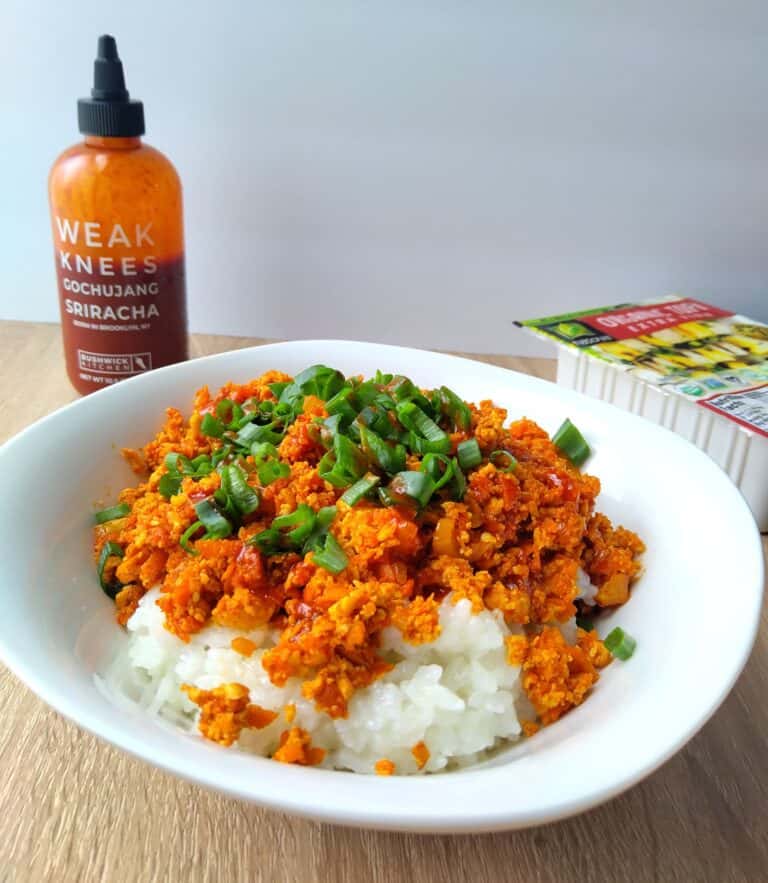 a bowl of bright red grown tofu with rice, and gochujang sriracha and a tofu pack behind it