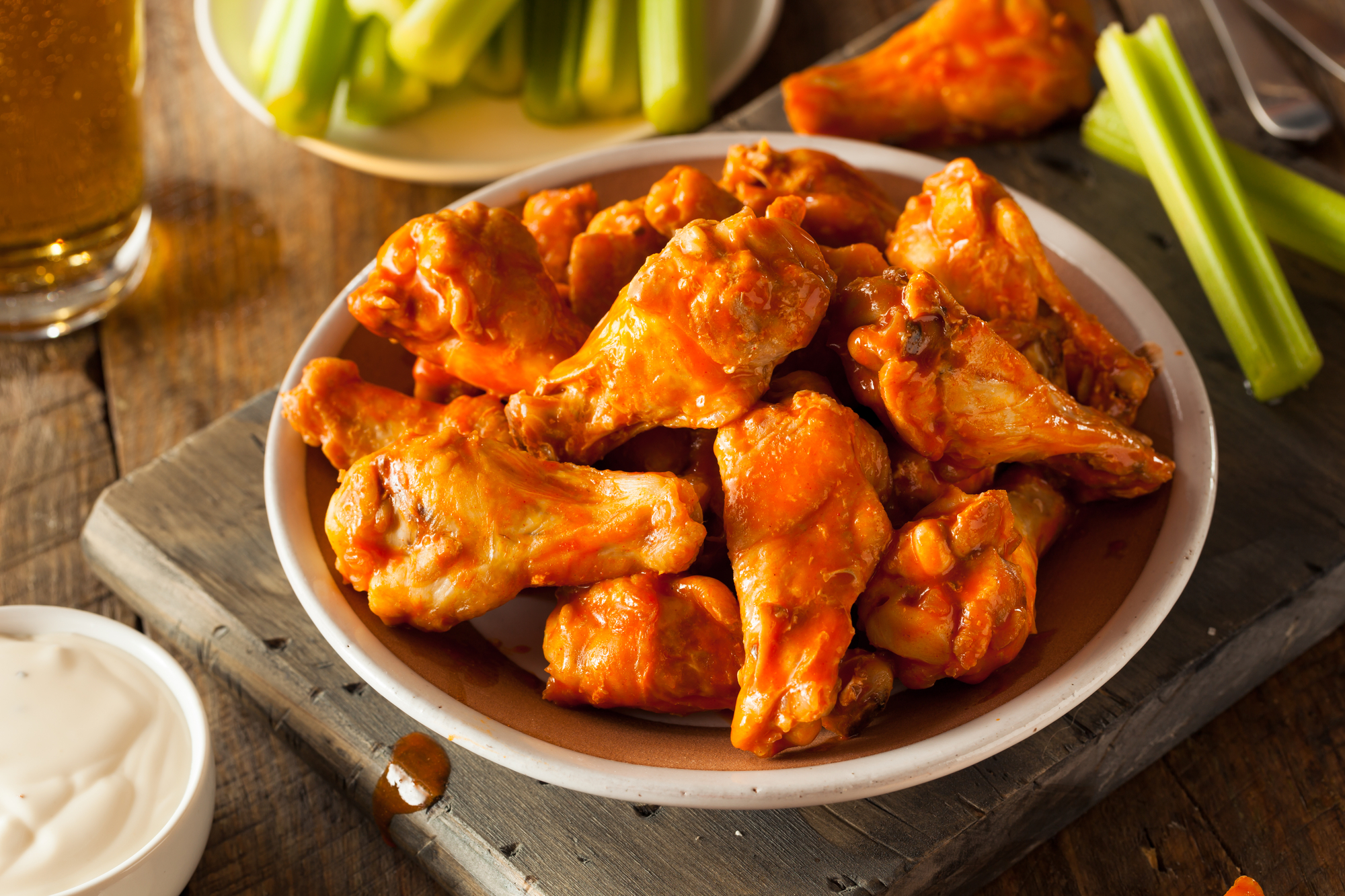 a platter of buffalo wings surrounded by celery, a dish of creamy dressing, and a glass of beer.